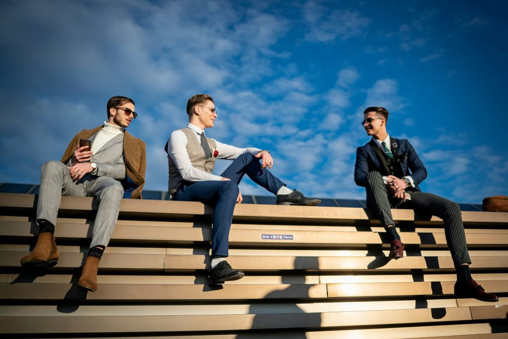 An image showing three stylish men in tailored outfits sitting on a rooftop ledge under a bright blue sky.