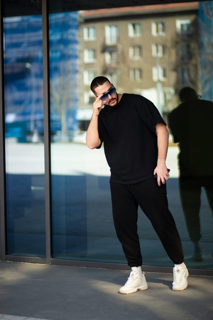 Image showing a man in a relaxed black T-shirt and joggers with white Fila sneakers and blue sunglasses posing near reflective glass windows.