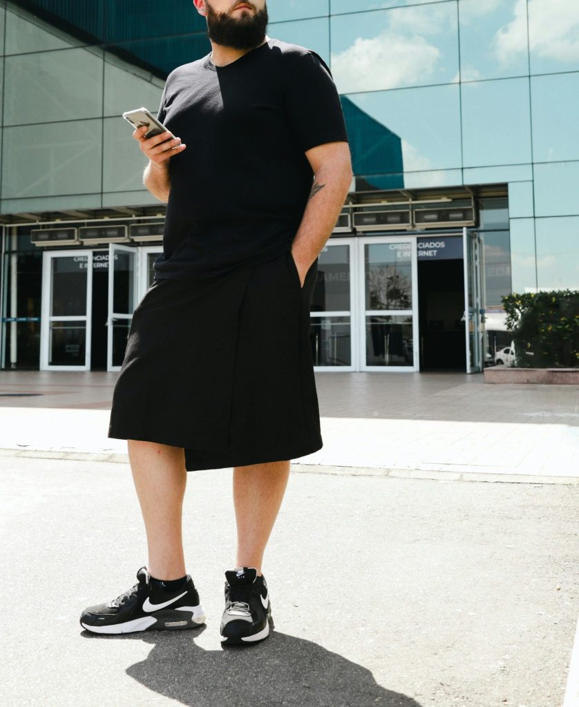 Image showing a bearded man wearing an oversized black T-shirt, long black shorts, and Nike sneakers standing outside a modern glass building.