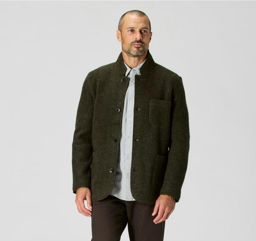 An image showing a man wearing a dark olive chore coat layered over a light gray button-down shirt, styled in a clean and minimal outfit.