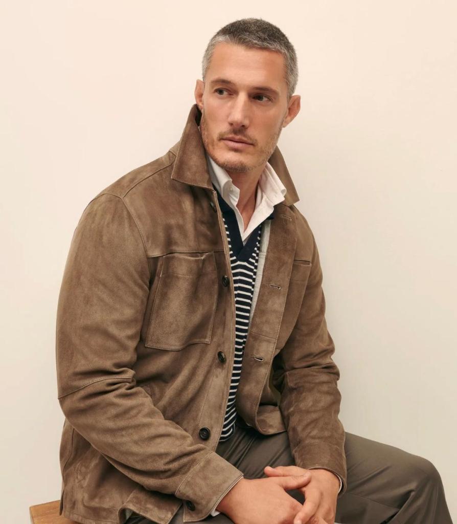 An image showing a man wearing a taupe suede shirt jacket over a striped sweater and white button-down, styled in a refined layered outfit.