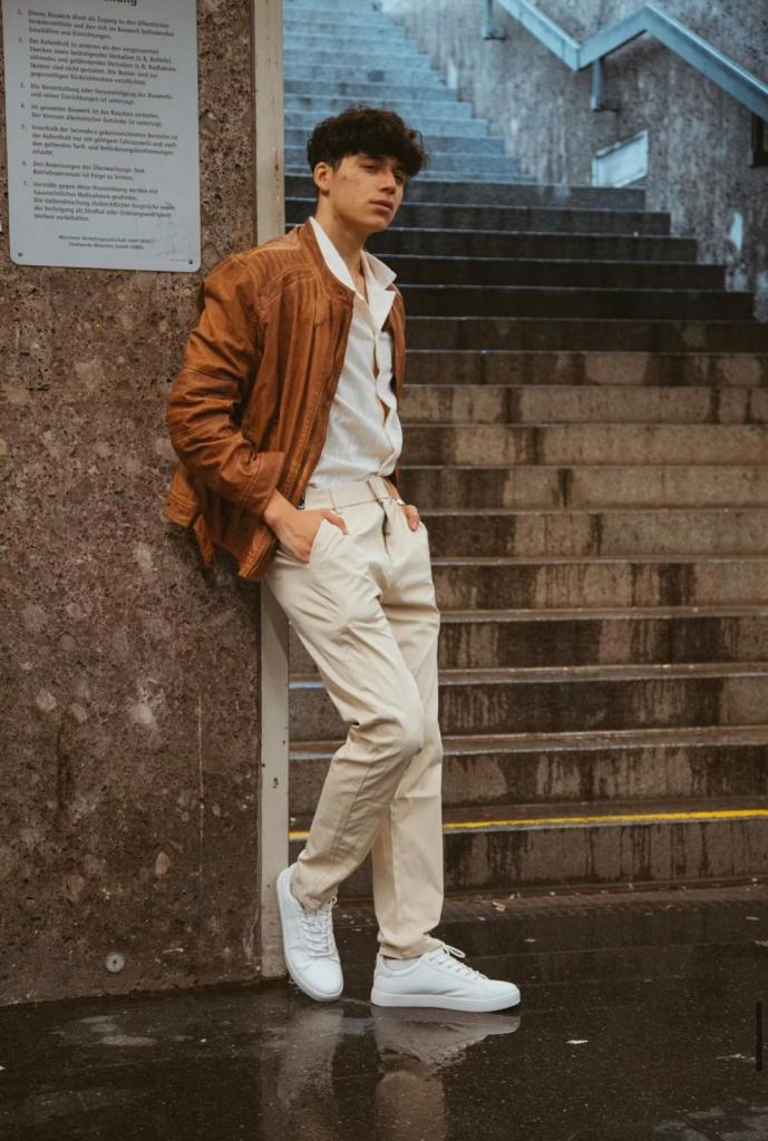 An image showing a man leaning against a stone wall near a staircase, wearing a brown leather jacket over a white shirt, cream pleated trousers, and white sneakers. His hands are in his pockets, and the wet pavement reflects his shoes.