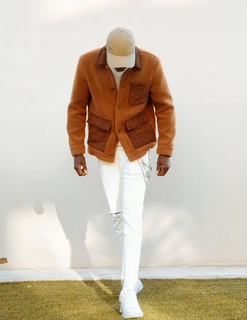 An image showing a man wearing a camel-brown fleece jacket with darker brown pockets over a white shirt, paired with white ripped jeans, a beige baseball cap, and white sneakers. He is looking down while walking outdoors on grass against a plain light-colored wall.