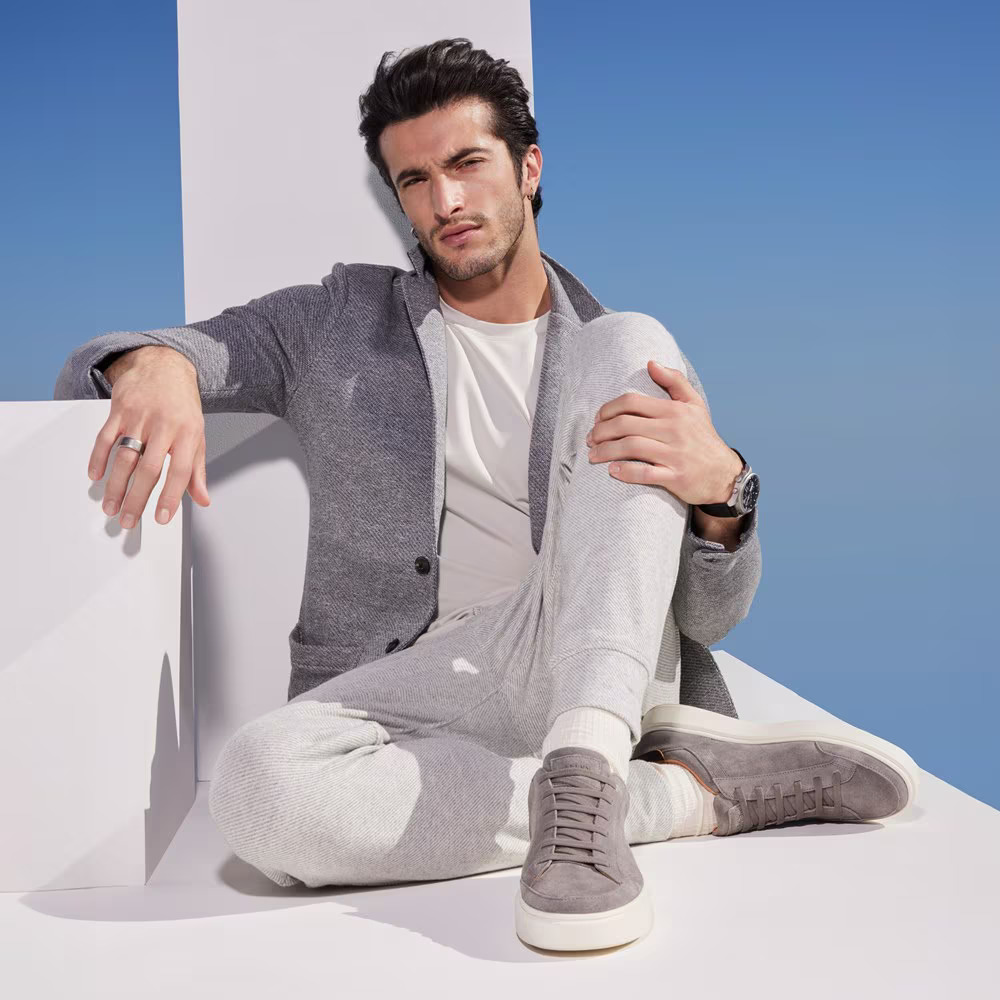 Image showing a stylish man wearing a grey blazer, white t-shirt, grey joggers, and grey suede sneakers against a blue sky background