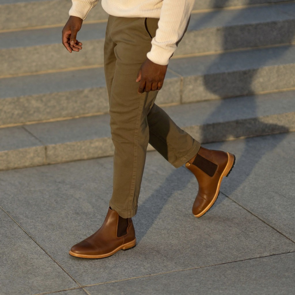 Image showing a man walking up stone steps wearing olive chinos, a cream sweater, and brown leather Chelsea boots
